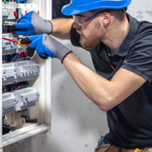 La imagen muestra a un técnico o ingeniero eléctrico trabajando en un panel eléctrico industrial. El profesional está equipado con medidas de seguridad, como un casco azul, gafas protectoras y guantes de trabajo. Está utilizando un destornillador para ajustar o conectar componentes en el panel, que contiene diversos dispositivos eléctricos como breakers y cables organizados.